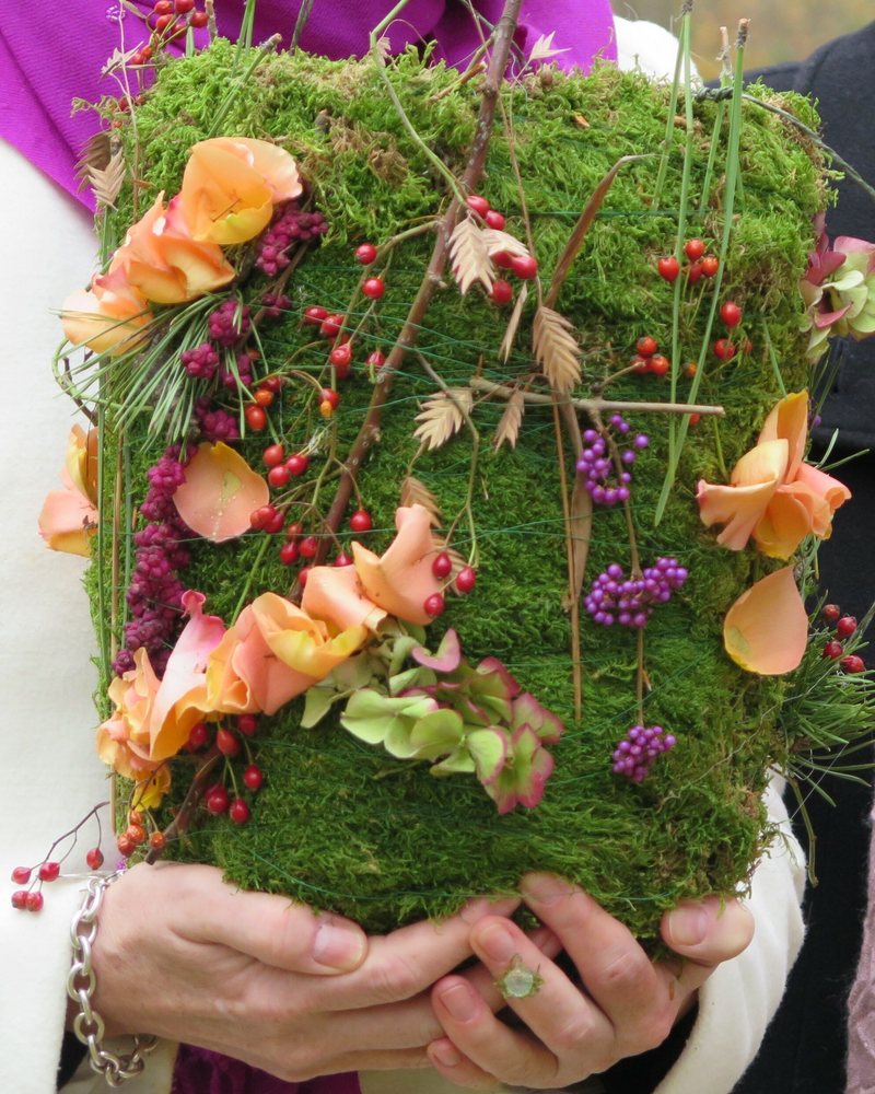 Hände halten eine mit Blumen und Beeren geschmückte Urne aus Moos
