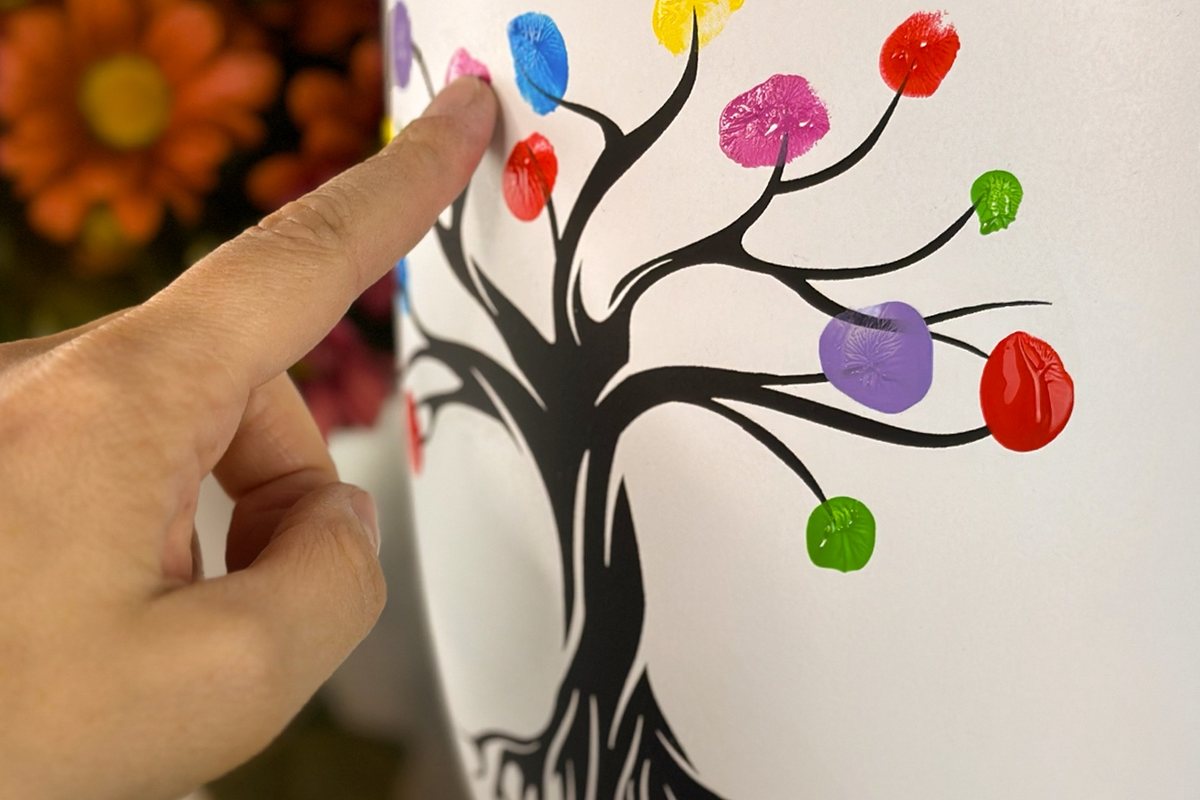 Ein Finger setzt einen bunten Farbabdruck an einen gemalten Baum auf einer weißen Urne